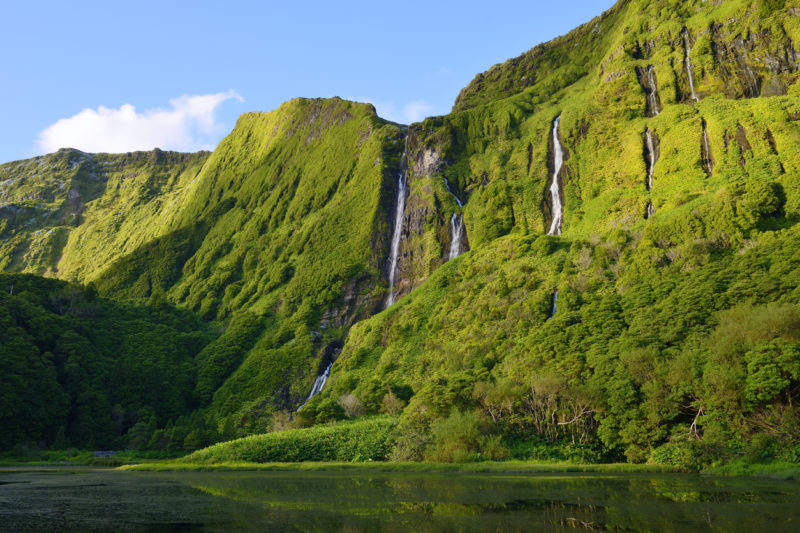 Poço da Alagoinha, Flores - Waterfalls on the island of Flores in Portugal. The image is giant mossy-green cliffs with waterfalls flowing down and a bright blue sky. 