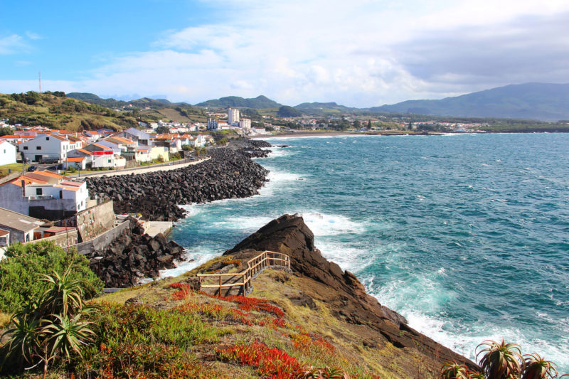 Sao Miguel Azores Portugal - a rocky coastline with white houses and a road next to the blue ocean and waves crashing, mountains in the distance. 