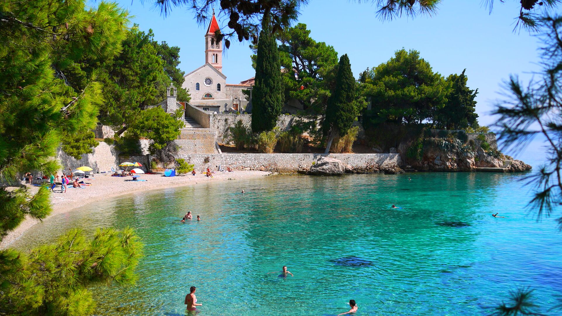 People swimming in turquoise waters in the ocean, with a beach and trees in Hvar