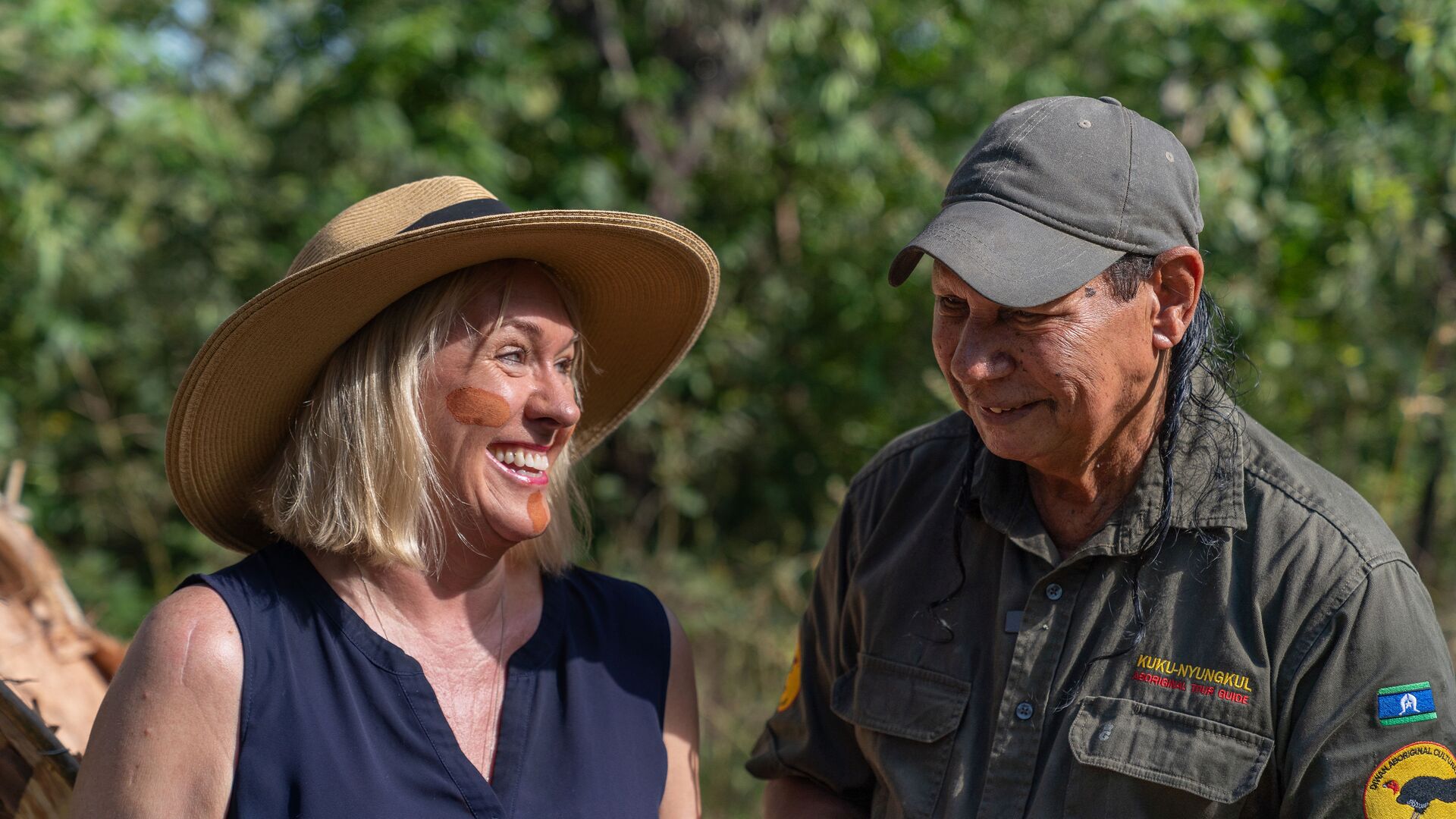 A local Aboriginal man, Mook, smiles next to a traveller with traditional face paint during a smoking ceremony.