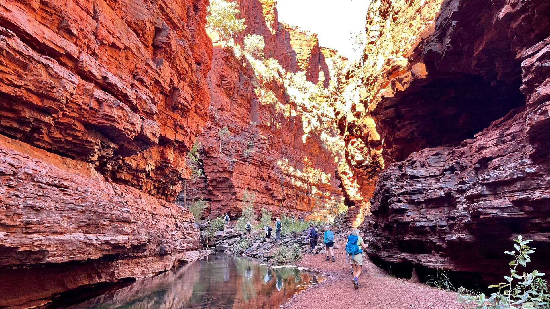 People walk along a track next to orange rocky cliffs in Karijini National Park.