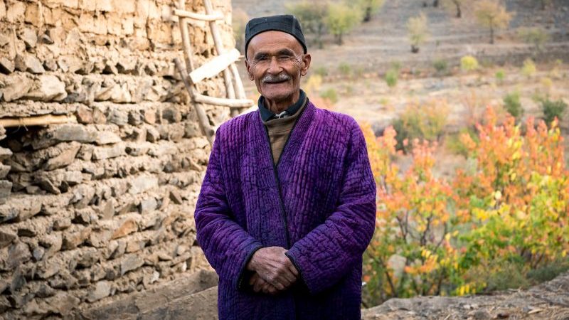 Local Uzbek man in traditional purple dress stands outside his house and poses for a photo