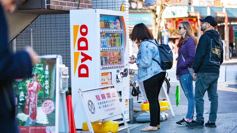 Woman stands in front of a drinks vending machine on a busy street in Japan.