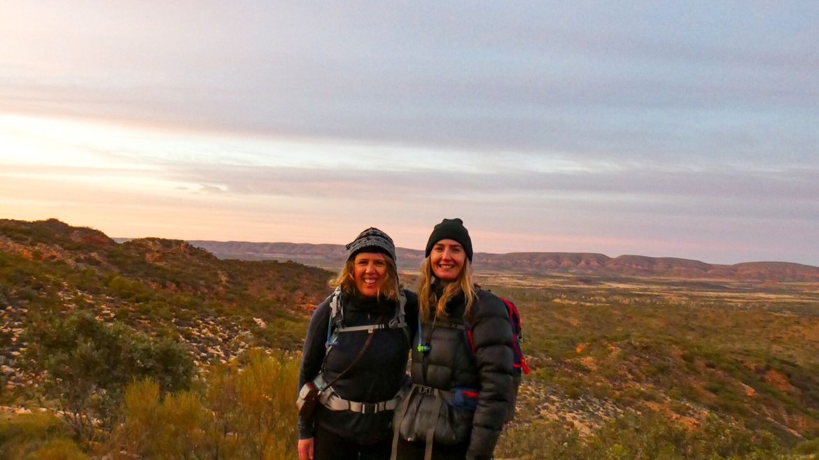 Two women smile with a sunset over the Larapinta Trail behind them.