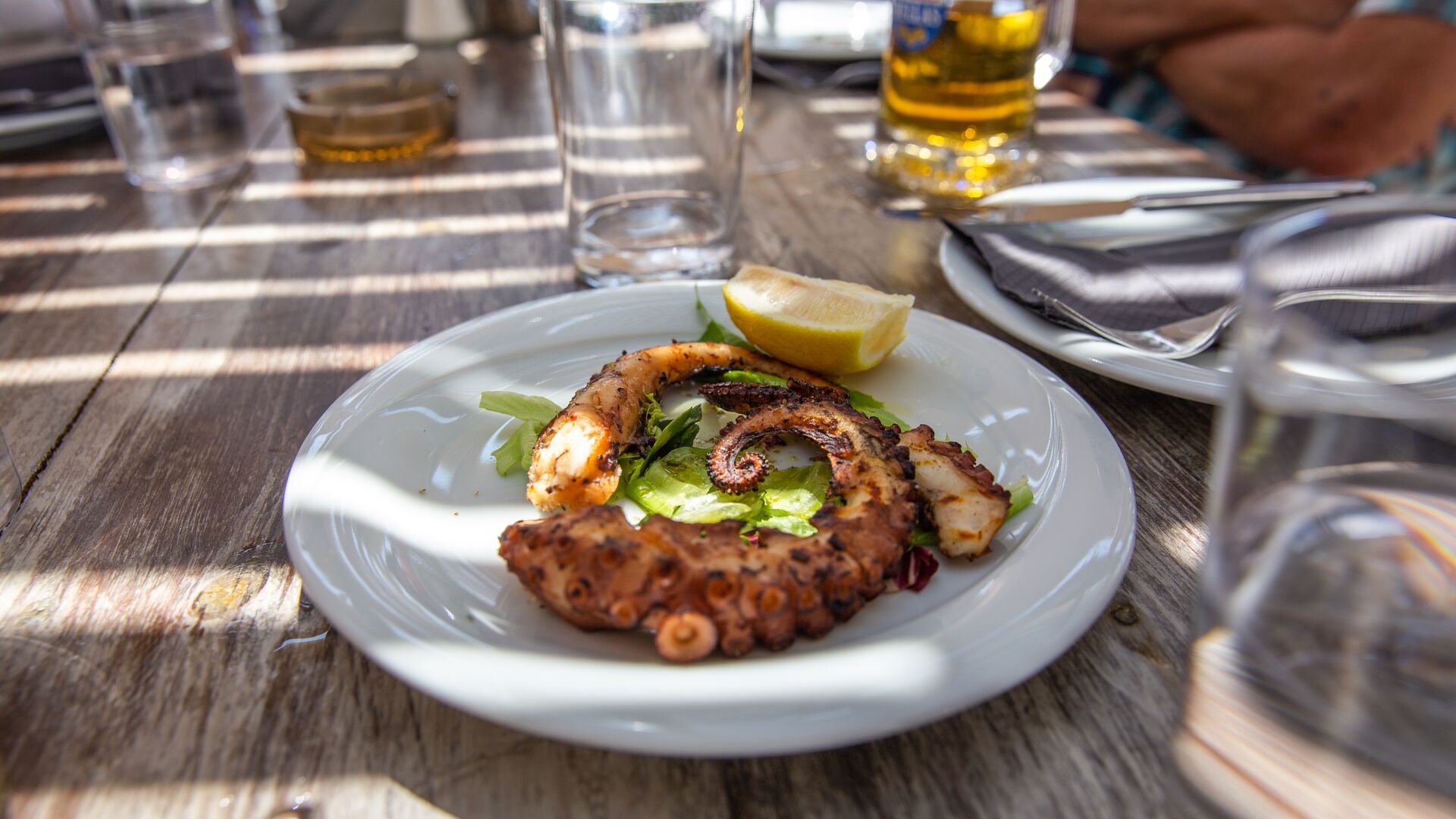A chargrilled octopus tentacle sits on a white plate with a lemon wedge. Also on the table is a glass of beer.