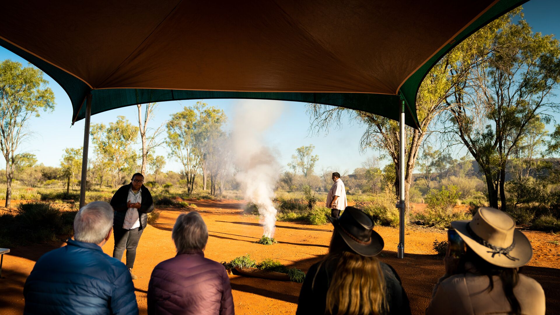 Four travellers sat on a bench while two Aborignal guides explain what a smoking ceremony is.