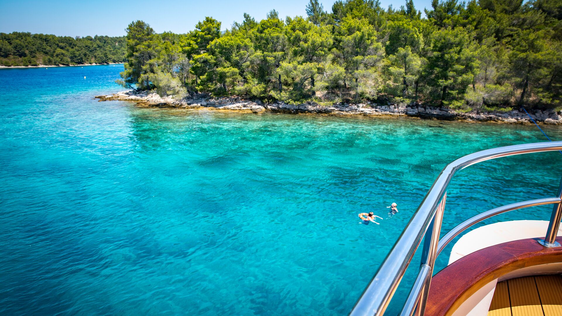Two travellers swim in turquoise waters in front of a boat with a brown deck and silver railings. An island covered in green trees is in the distance.