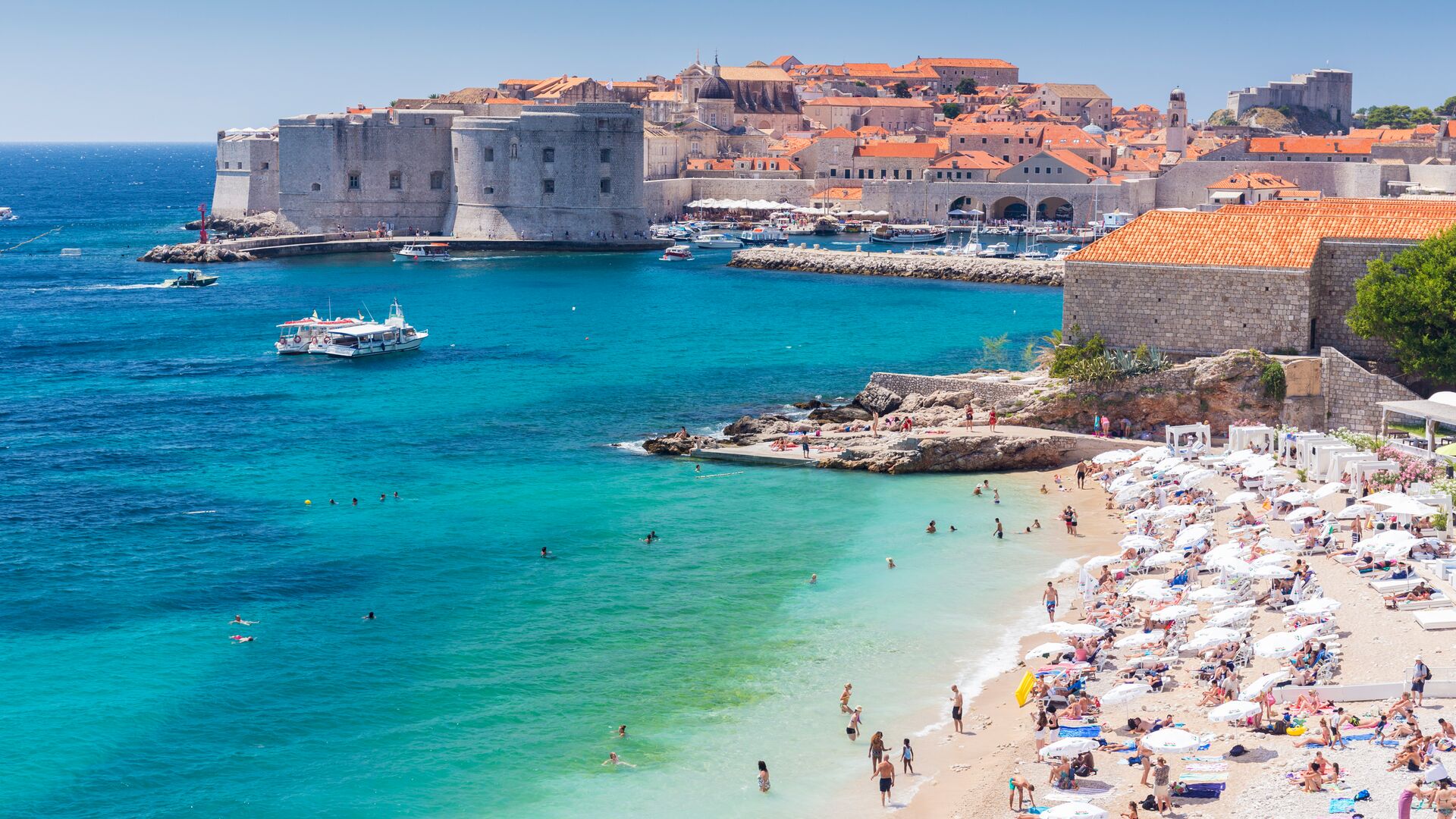 The walled city of Dubrovnik is in the background of the image. The roofs of the buildings are orange. In the foreground, a beach is full of people. The sea surrounding the city is bright turquoise.