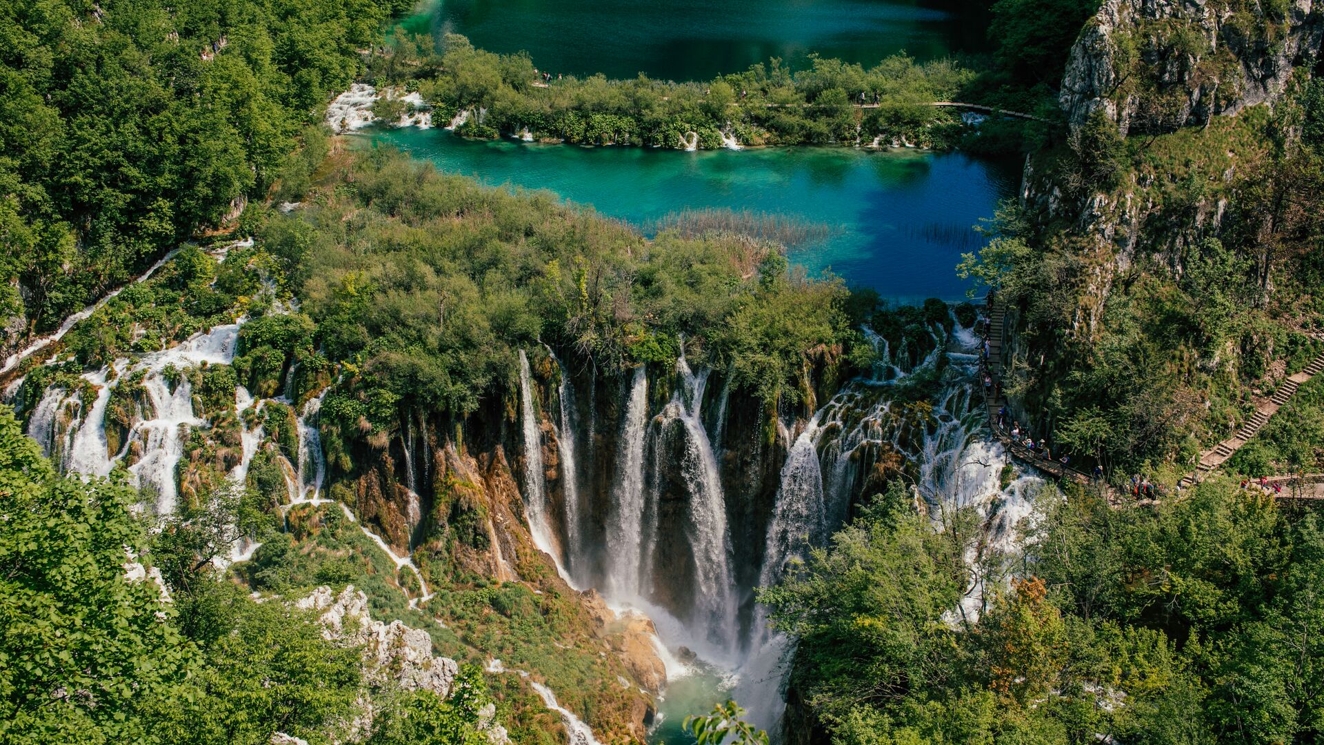 An aerial view of Plitvice National Park, Croatia. 15 waterfalls are visible in a deep green forest. In the distance, a bright blue pool feeds the falls.