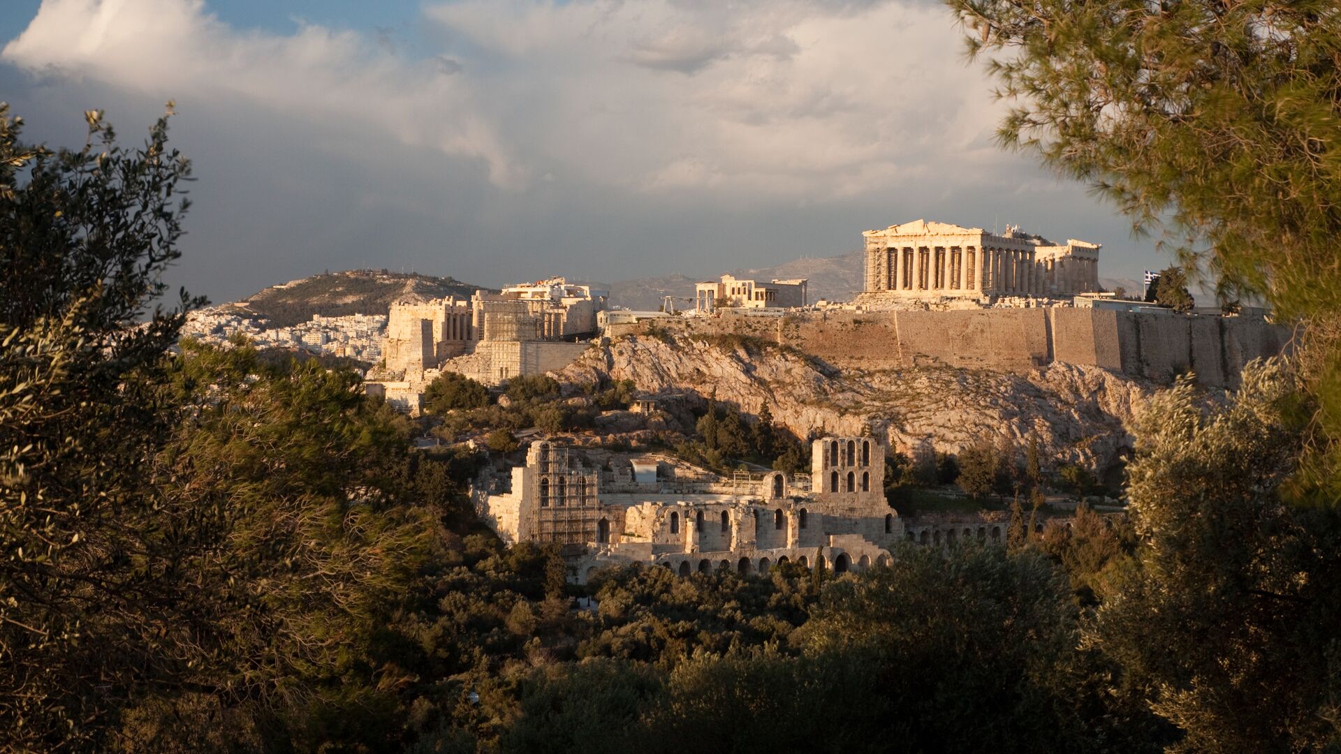 The ruins of the Acropolis in Athens are perched high above the city. The sky is a moody gray and the light on the buildings is golden. Trees frame the foreground of the image.