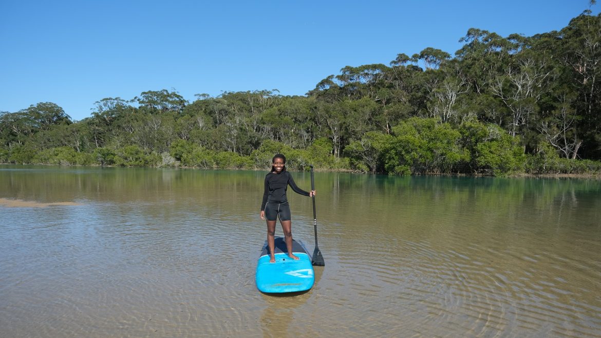 A stand up paddle boarder smiles for a photo on a quite creek