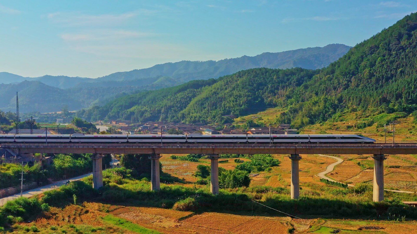 A high speed train crosses a bridge in rural China, passing terraced rice paddies and forested mountains.