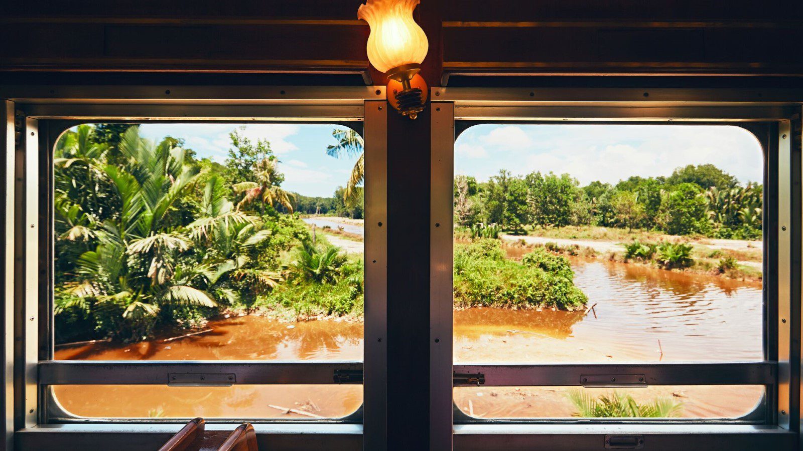 A view from the inside of a train carriage, looking out over the Malaysian countryside, including a wide river and palm trees.