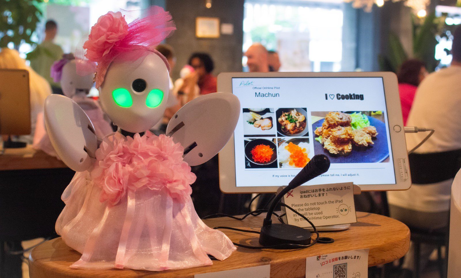 A tiny robot in a pink dress with lit-up green eyes, infront of a tablet and a microphone.