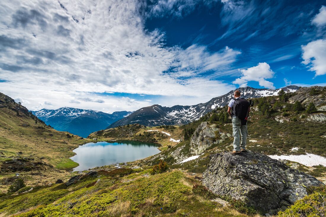 A hiker stands with their back to the camera on a rock while gazing over a small lake, with mountains in the background