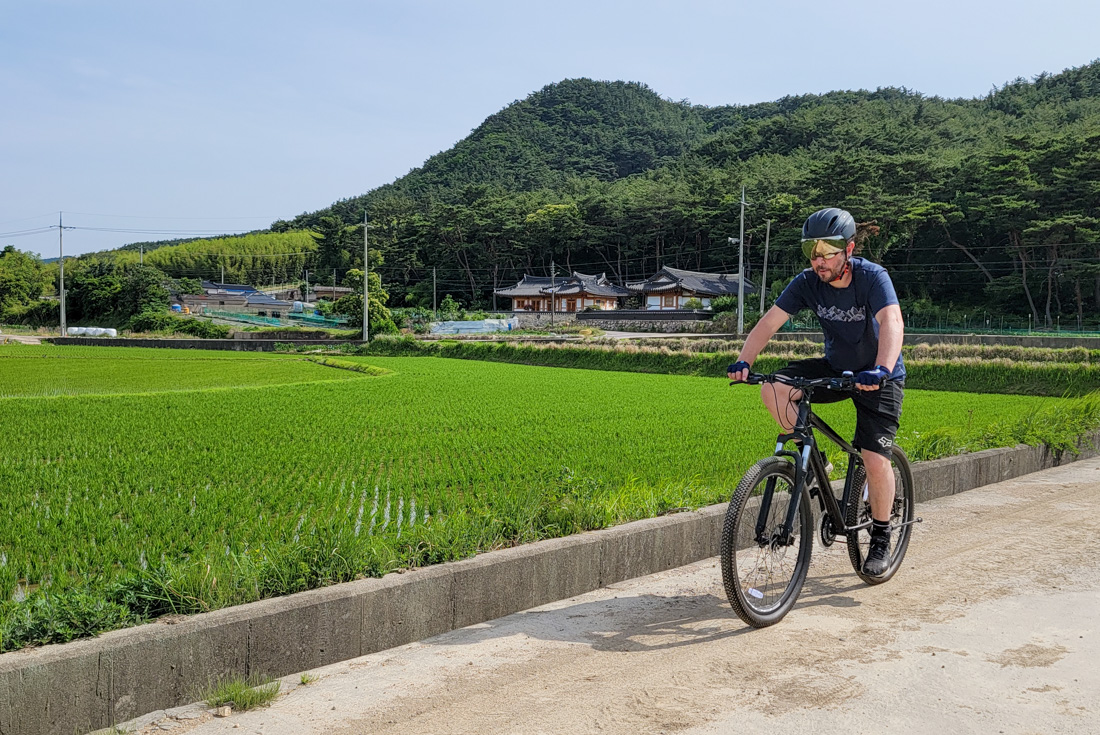 Cyclist in helmet and gear cycling in rural South Korea past traditional houses and fields