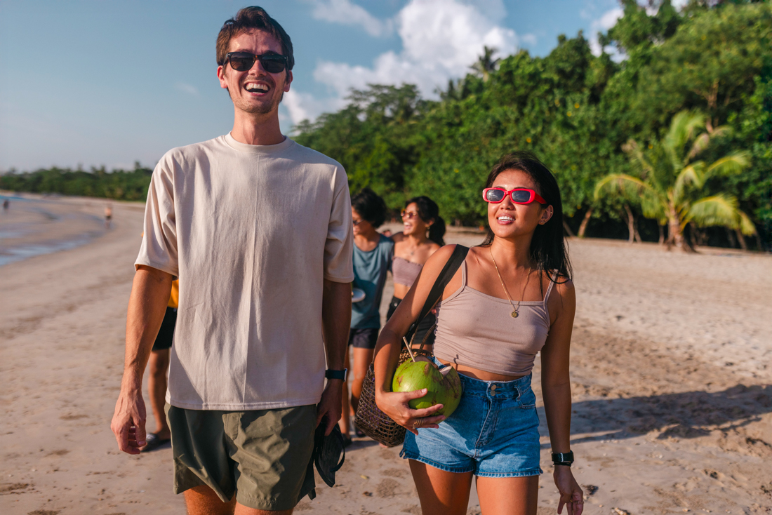Young Intrepid travellers stroll along a beach in the Philippines with drinking coconuts laughing and enjoyng the sun