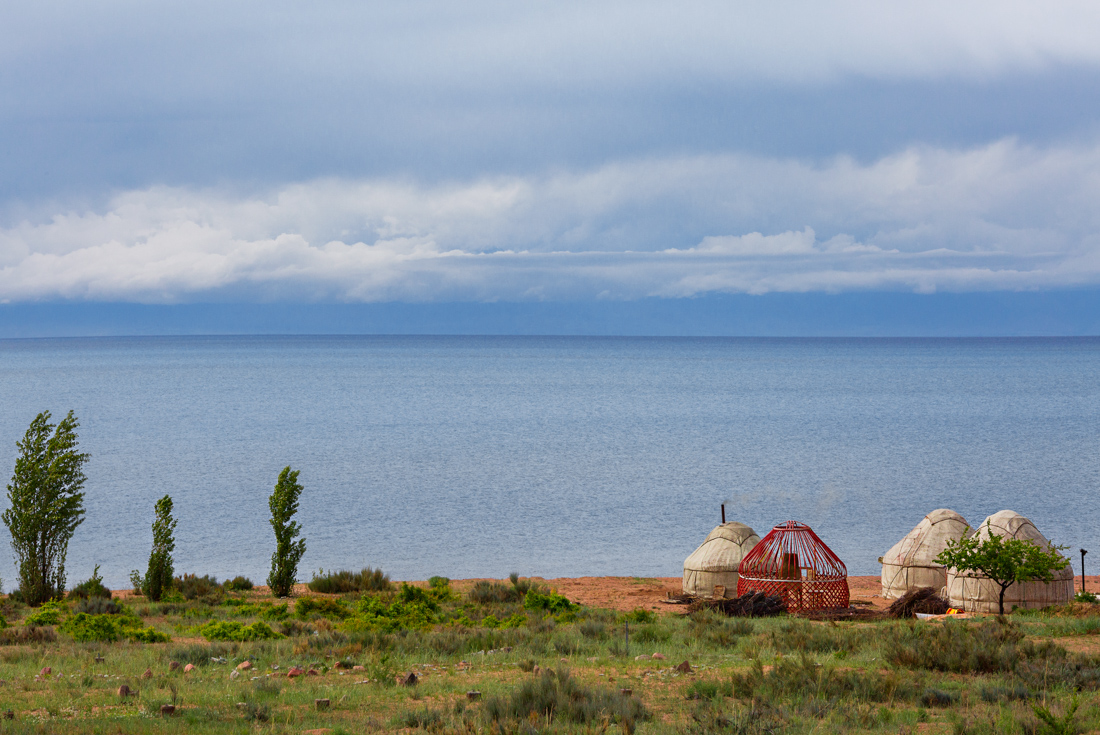A set of yurts with one partially assembled sit on above the shore of Issy-Kul Lake with mountains in the distance