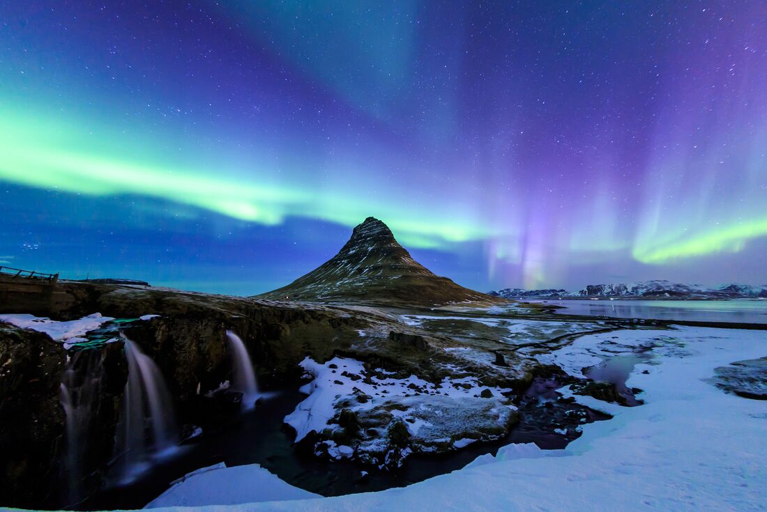 A wide shot of Kirkjufell hill with blue, purple and green aurora lights illuminating the sky